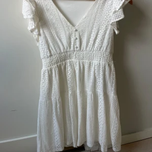 Vit spetsklänning från Vero Moda - Supersöt vit kortklänning från Vero Moda med v-ringning, volangärmar och smock i midjan. Klänningen har spetsdetaljer och är perfekt för sommarens alla tillfällen. Lätt och luftig med romantisk känsla.