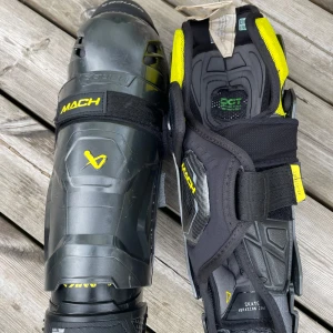 Bauer Mach hockeyskydd/benskydd  - Säljer ett par Bauer Mach hockeyskydd i svart och gul design. De har robusta skyddszoner, justerbara kardborreband och ventilerande meshdetaljer. Perfekta för dig som vill ha grymt skydd och cool look på isen.