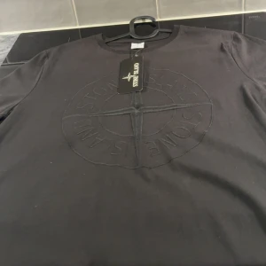 Svart Stone Island t-shirt med brodyr - En helt ny och oanvänd Svart t-shirt från Stone Island med stort broderat logomärke framtill. Klassisk rund hals och korta ärmar. Tillverkad i 100% bomull för skön känsla och snygg passform. Perfekt för dig som gillar stilrena streetwear-plagg med exklusiv känsla.