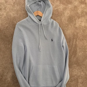 Ljusblå hoodie från Polo Ralph Lauren - Snygg ljusblå hoodie från Polo Ralph Lauren med broderad logga på bröstet. Tröjan har huva med snörning, känguruficka och är stickad i bomull. Perfekt för en chill och stilren look.