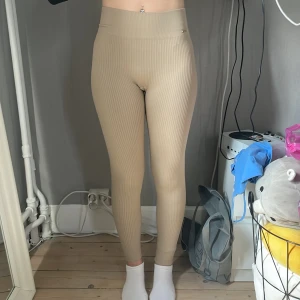 Beige ribbade leggings Ellen New York - Snygga beige ribbade leggings från Ellen New York med hög midja och tight passform. De är stretchiga och har diskret logga bak samt en liten metallbricka framtill. Perfekta för en chill och trendig look.
