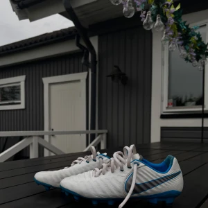 Nike Tiempo vita fotbollsskor - Nike Tiempo fotbollsskor i vitt med blå detaljer och silverfärgad swoosh. Skorna har snörning, rund tå och platt sula med dobbar för bra grepp på planen. Ovandelen är i syntetmaterial och insidan är fodrad i blått. Perfekta för dig som vill sticka ut på fotbollsplanen. En gräsfläck på baksidan men inget man tänker på, skriv för fler bilder. (Pris kan diskuteras)