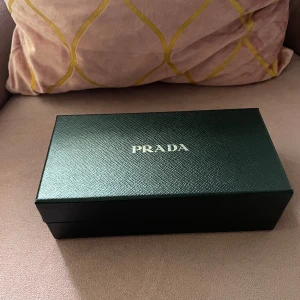 Svart plånbok från Prada i skinn - Snygg svart plånbok från Prada i äkta skinn med klassisk trekantslogga i metall framtill. Plånboken har ett stilrent kuvertformat lock och levereras i originalask med tillhörande certifikat. Perfekt accessoar för dig som gillar lyxiga detaljer.