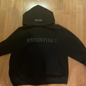 Svart Essentials hoodie från Fear of God - Svart hoodie från Fear of God Essentials med stor logga på bröstet och detaljer på huvan och ärmen. Klassisk känguruficka framtill och oversized passform. Perfekt för dig som gillar streetwear och vill ha en stilren look. Tröjan är Storlek M och kan passa L eftersom den är rätt så oversized i passformen