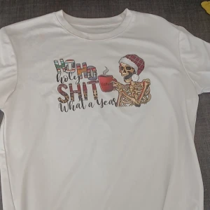 Vit jul-tshirt med skelettmotiv XS - Vit t-shirt i polyester med tryck av ett skelett i tomteluva som håller en kaffemugg och texten 'Ho Ho Holy Shit What a Year'. Perfekt för dig som gillar humor och juliga motiv.