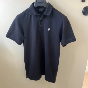 Svart pikétröja från Sunwill - Klassisk svart pikétröja från Sunwill med korta ärmar och krage. Tröjan har två knappar framtill och en liten vit logga på bröstet. Tillverkad i mjuk bomull som känns skön mot huden. Perfekt för en clean och stilren look.