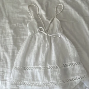 Vit somrig tröja med spets - Superfin vit kortklänning med tunna axelband, v-ringning och knyt framtill. Klänningen har broderade spetsdetaljer och är gjord i ett lätt, luftigt material som passar perfekt för varma dagar.