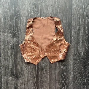 Bronsfärgad paljettväst - Glittrig väst i brons med tättsydda paljetter över hela framsidan. Modellen är ärmlös med djup v-ringning och hakar som knäppning framtill. Perfekt för att sticka ut och ge extra bling till din outfit.