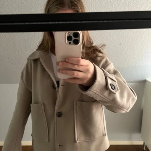 Beige ulljacka från Nelly, stl S - Snygg beige jacka från Nelly i ullmix med klassisk krage, stora fickor framtill och knappar. Jackan har långa ärmar med knappdetalj vid mudden och en rak, lite boxig passform. Aldrig använd men har hängt i garderoben ett år