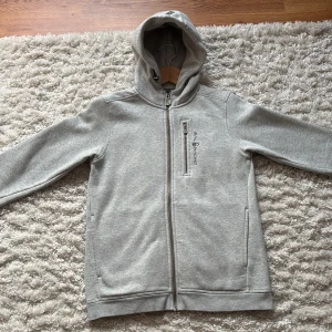 Grå hoodie från Sail Racing - Snygg ljusgrå hoodie från Sail Racing med dragkedja hela vägen, huva och bröstficka med dragkedja. Broderad logga på bröstet och en stilren look. Perfekt för lager-på-lager under höst och vinter.