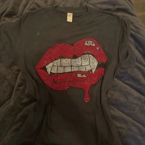 Svart t-shirt med glittriga röda läppar - Cool svart t-shirt från Gildan med ett stort tryck av glittriga röda läppar och vita tänder framtill. T-shirten är kortärmad och gjord i mjuk bomull, perfekt för att sticka ut i mängden. Snygg och edgy vibe!