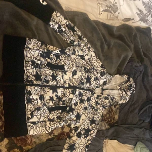 Blommig hoodie med text Queen - Snygg hoodie med svartvita blommor och stjärnor, dragkedja framtill och huva. Svarta muddar och texten 'QUEEN' tryckt i vitt på ena ärmen. Perfekt statement-plagg för dig som vill sticka ut.
