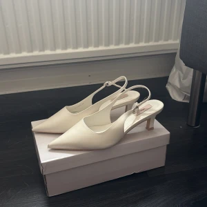 Vita låga klackskor  från Nelly - Stilrena vita klackskor från Nelly med spetsig tå och smal rem runt hälen. Skorna har en låg klack och är tillverkade i syntetmaterial med en elegant, minimalistisk look. Perfekta för dig som gillar klassisk och clean stil.