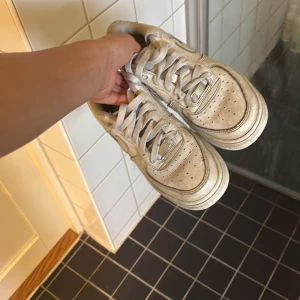 Nike Air Force 1 vita sneakers - Klassiska Nike Air Force 1 sneakers i vitt skinn med perforerad tå och snörning. Modellen har en chunky sula och den ikoniska swoosh-loggan på sidan. Perfekta för dig som gillar streetstyle och tidlös design.