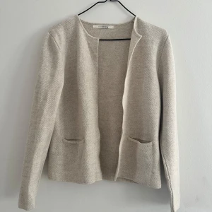 Beige stickad cardigan jacka Sibin Linnebjerg - Snygg beige stickad cardigan jacka från Sibin Linnebjerg. Rak modell med öppen front och två fickor framtill. Mjuk och skön struktur, perfekt att slänga över en topp. Enkel och stilren design som funkar till det mesta.