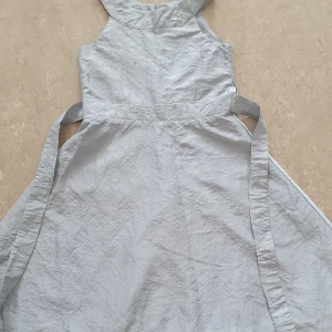 Klänning - Supersöt ljusgrå klänning från Lindex med broderade detaljer och små paljetter i midjan. Klänningen har knytband i sidorna, dragkedja bak och är ärmlös. Perfekt för dig som gillar en stilren look med lite extra bling.
