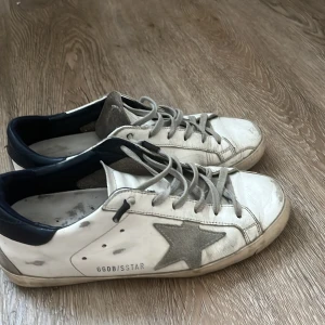 Golden Goose vita sneakers med stjärna - Golden Goose sneakers i vitt skinn med grå mockadetaljer och ikonisk stjärna på sidan. Skorna har grå snörning, mörkblå hälkappa och platt sula. Perfekta för dig som gillar en avslappnad och trendig look.