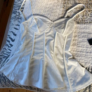 Vit satin korsettopp från hm - Supersnygg vit korsettopp i satin från hm med tunna axelband och markerade sömmar framtill. Toppen har utsvängd nederdel och smock i ryggen för extra komfort. Perfekt att styla med jeans eller kjol för en trendig look.