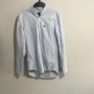 Ljusblå skjorta från Lyle & Scott S - Snygg ljusblå skjorta från Lyle & Scott i storlek S. Klassisk modell med button down-krage, lång ärm och gul broderad logga på bröstet. Skjortan är tillverkad i bomull och har knappar framtill. Perfekt för en clean och stilren look.