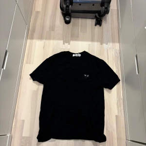 Svart Comme des Garçons Play t-shirt - Svart t-shirt från Comme des Garçons Play med klassisk rund hals och korta ärmar. På bröstet sitter det ikoniska hjärtat med ögon i svart. T-shirten är i mjuk bomull och har en clean, stilren vibe som passar till allt. Typ aldrig använd storlek L men sitter som en mindre M. Pris kan diskuteras 