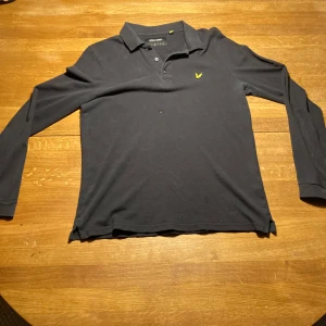 Svart långärmad pikétröja Lyle & Scott - Svart långärmad pikétröja från Lyle & Scott i 100% bomull. Klassisk krage med knappar och den ikoniska gula logon broderad på bröstet. Perfekt för dig som gillar stilren och enkel design med en sportig touch.