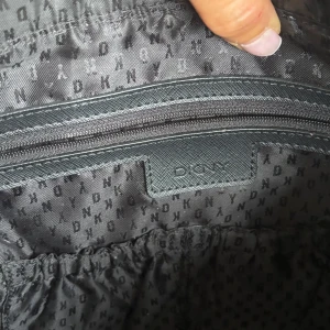 Svart axelväska från DKNY med kedjedetaljer - Snygg svart axelväska från DKNY i skinn med guldfärgade kedjor och logga framtill. Väskan har dragkedja upptill, innerfack och ett dekorativt DKNY-hänge. Perfekt storlek för att få plats med allt du behöver och ger en lyxig känsla till din outfit.