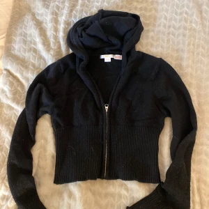 Svart croppad hoodie från Gina Tricot - Svart croppad hoodie från Gina Tricot med dragkedja framtill och bred ribbad mudd i midjan. Tröjan har långa ärmar och huva, perfekt för en chill och stilren look. 