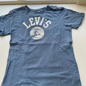 Blå Levi's t-shirt med surftryck - Snygg blå t-shirt från Levi's med vit logga och surfmotiv på bröstet. Klassisk passform med korta ärmar och rund hals. Tillverkad i 100% ekologisk bomull, perfekt för en chill och avslappnad stil.