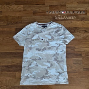 Beige Tommy Hilfiger T-Shirt - Tommy Hilfiger T-shirt i storlek S, i nyskick! Snygg beige/vit camo-mönstrad design med broderad Tommy Hilfiger-logga framtill (EST. 1985). Mjuk och skön bomullsblandning, perfekt för vardagsbruk eller streetwear. Aldrig använd utomhus, inga fläckar eller slitage. Skickas snabbt vid köp! ✨👕📦