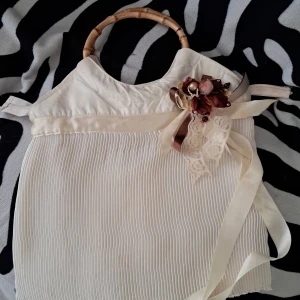 Vintage Vit handväska med bambuhandtag och rosett - Unik vit handväska med bambuhandtag och dekorativ rosett i spets och satinband. Väskan har plisserad framsida (några fläckar) och en vacker blomdekoration i brunt och pärlor. Perfekt accessoar för dig som gillar vintagevibbar och romantiska detaljer. Lite smutsig inuti, men går nog att torka bort :) 