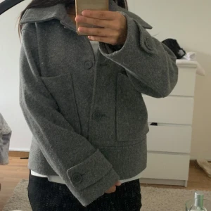 Grå ulljacka med stora knappar (kappa)  - Säljer en grå, kortare jacka i ull med stora fickor framtill och bred krage. Jackan har stora knappar och en boxig, loose passform som ger en chill vibe. Perfekt för lager-på-lager under höst och vinter. Enkel att styla till både jeans och kjol.