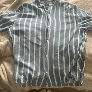 Blåvit randig skjorta från H&M - Snygg blå skjorta med vita vertikala ränder från H&M. Skjortan har klassisk krage, knäppning framtill och en relaxed passform. Perfekt för dig som gillar en stilren och fräsch look. Materialet känns lätt och passar bra till varmare dagar.