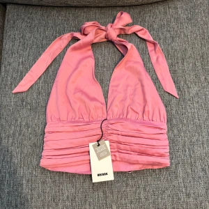 Rosa halterneck topp från Bik Bok - Supersöt rosa halterneck topp från Bik Bok i glansig polyester. Toppen har breda knytband i nacken, rynkade detaljer och små klädda knappar framtill. Perfekt för dig som vill sticka ut med en feminin och trendig look.