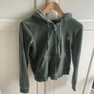 Grön velour hoodie Juicy Couture S - Mörkgrön velour hoodie från Juicy Couture med glittrande JC-logga på bröstet och stor Juicy-text i strass på ryggen. Tröjan har huva, dragkedja med J-berlock, känguruficka och långa ärmar. Perfekt för dig som gillar 00-talsvibbar och comfy stil.