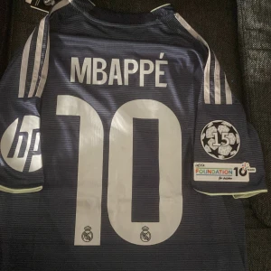 Real Madrid Mbappé fotbollströja  - Snygg Real Madrid fotbollströja med Mbappé och nummer 10 på ryggen. Mörkblå med tunna horisontella ränder, vita detaljer och klubbmärke broderat på bröstet. Adidas-logga, HP-sponsor och Champions League 15-patch på ärmen. Tillverkad i lätt funktionsmaterial. Storlek M