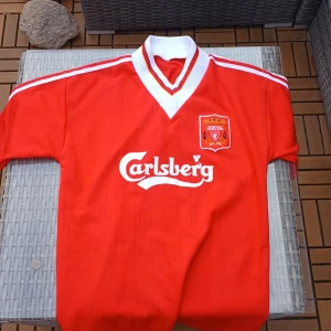 Liverpool retro fotbollströja Carlsberg - Klassisk röd Liverpool fotbollströja med Carlsberg-tryck på bröstet och klubbmärke broderat på vänster sida. Tröjan har vit V-ringad krage med röda detaljer och vita ränder på axlarna. Tillverkad i lätt och ventilerande material, perfekt för match eller street wear.