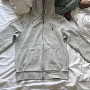 Grå hoodie från Polo Ralph Lauren - Snygg ljusgrå hoodie från Polo Ralph Lauren med dragkedja, huva med snörning och klassisk logga på bröstet. Tillverkad i mjuk bomullsmix, perfekt för chill dagar. Fickor framtill och relaxed fit för en casual vibe.