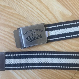 Randigt bälte från Hampton Republic - Snyggt randigt bälte från Hampton Republic med metallspänne där det står 'Classics 27'. Bältet är svart och vitt med breda ränder och har en sportig känsla. Perfekt accessoar för att lyfta din outfit.