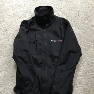 Svart Sail Racing Gore-Tex vindjacka - Svart vindjacka från Sail Racing i Gore-Tex-material. Jackan har hög krage, hel dragkedja framtill och diskret logga på bröstet. Perfekt för dig som vill ha en stilren och funktionell jacka med sportig känsla.