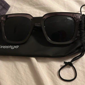 Svarta solglasögon från Prestige - Snygga svarta solglasögon från Prestige med bred, fyrkantig båge i transparent plast. Glasen är mörka och ger en cool look. Kommer med svart Prestige-fodral. Perfekta för att lyfta din stil i sommar.