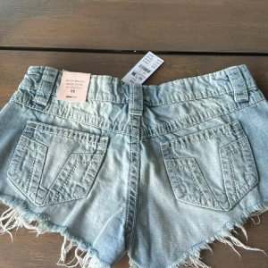 Ljusblåa jeansshorts från Gina Tricot - Snygga ljusblå jeansshorts från Gina Tricot med låg midja, slitna detaljer och fransig kant. Klassisk femficksmodell och bälteshällor. Perfekta för sommaren och ger en avslappnad vibe till din outfit.