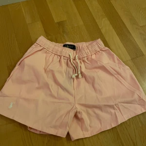 Ljusrosa shorts från Ralph Lauren - Snygga ljusrosa shorts från Ralph Lauren med elastisk midja och vit snörning framtill. Klassisk broderad logga i vitt på ena benet. Tillverkade i mjuk bomull, perfekt för varma sommardagar. Avslappnad passform och stilren design.