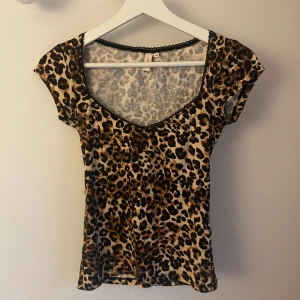 Leopardmönstrad topp från Nelly 🐆 - Snygg leopardmönstrad topp från Nelly i storlek XS. Toppen har korta ärmar, v-ringning med svart spetskant och är figurnära. Perfekt för dig som vill sticka ut med ett djurmönster och cool vibe.