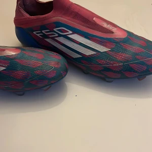 Adidas F50 fotbollsskor blå/röda - Säljer ett par Adidas F50 fotbollsskor i en riktigt snygg färgkombo av blått, rött och vitt. Skorna har ett mönstrat yttre med vita detaljer och klassiska Adidas-ränder. Ovandelen är i syntetmaterial och skon har platt sula med dobbar för gräsplan. Jag har tappat bort väskan har använt dom i ett tag nu men känns fortfarande som att dom är nya.