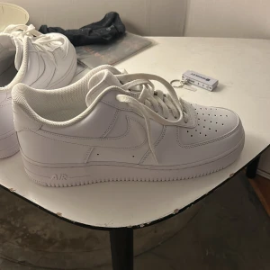 Nike Air Force 1 helvita sneakers storlek 46 (helt oanvända)  - Klassiska Nike Air Force 1 sneakers i helvitt skinn med perforerad tå, rund tå och platt sula. Snygg och tidlös modell med snörning och diskret Nike-logga på sidan och hälen. Perfekta för dig som vill ha en clean och stilren look.