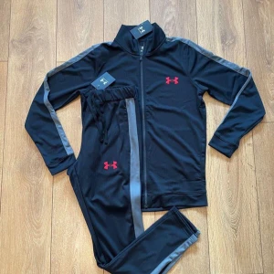 Under Armour tracksuit - Snyggt svart träningsset från Under Armour med gråa ränder längs ärmar och ben. Setet har dragkedja på jackan och röda loggor på både jacka och byxa. Perfekt för dig som gillar sportig stil och vill sticka ut lite extra på gymmet.