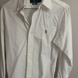 Vit skjorta från Ralph Lauren Slim Fit - Klassisk vit skjorta från Ralph Lauren i slim fit-modell. Skjortan har långa ärmar, knappar framtill och den ikoniska broderade polospelaren på bröstet. Tillverkad i mjuk bomull och passar perfekt till en stilren look.
