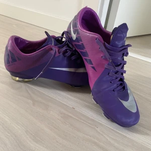 Lila Nike fotbollsskor strl 42 - Snygga fotbollsskor från Nike i lila och mörklila med silvriga detaljer och stora swoosh-loggor. Skorna har snörning och är tillverkade i syntetmaterial, med dobbar för bra grepp på planen. Perfekta för dig som vill sticka ut på matchen!