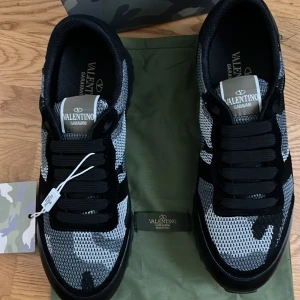 Valentino Garavani camo sneakers - Snygga Valentino Garavani sneakers med svart och grått camouflagemönster. Ovandel i mesh och mocka, svarta snören och robust sula med mönstrat grepp. Perfekta för dig som vill sticka ut med exklusiv streetstyle.