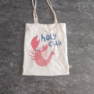 Vit tygkasse med röd krabba - Vit shoppingväska i bomull med tryck av en röd krabba och blå texten 'holy crab' på framsidan. Långa handtag gör den enkel att bära på axeln. Perfekt för att bära böcker, mat eller annat du behöver under dagen.